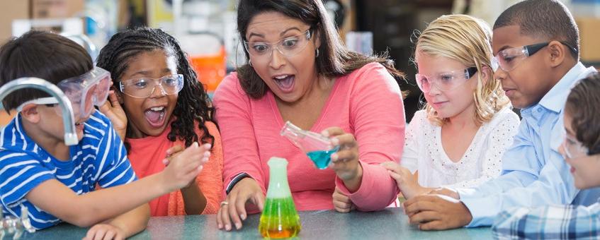 Teacher with students performing science experiment by combining different colored liquids in a beaker.